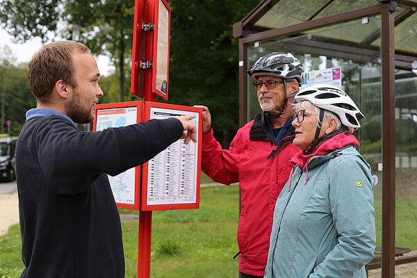 Ein Fachkraft im Fahrbetrieb erklärt den Fahrgästen an der Bushaltestelle den Fahrplan und die aktuellen Änderungen im Bezug auf den Fahrplanwechsel.