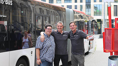 Drei Mobilitätsberaterkollegen stehen vor einem Linienbus und legen sich freundschaftlich die Arme auf die Schultern.