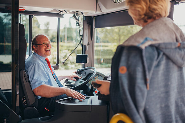 Busfahrer begrüßt lachend einen neu zugestiegenen Fahrgast.