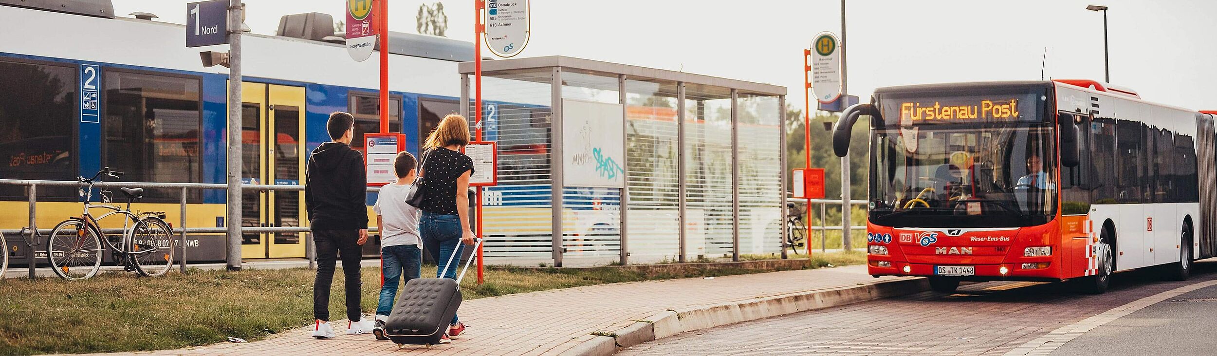 Personen mit Gepäckstück gehen vom Bahnsteig zum wartendem Bus an einer Haltestelle.