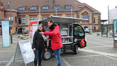 Ein Mobilitätsberater spricht vor dem Infomobil der V O S auf dem Bahnhofsvorplatz mit einer Passantin.