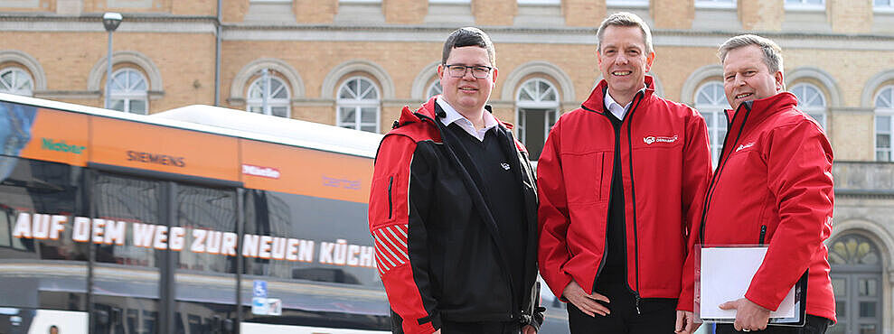 V O S Geschichte 2018. Drei Mobilitätsberater mit roten Jacken stehen vor einem Hintergrund mit Linienbus und dem Osnabrücker Landgericht. 