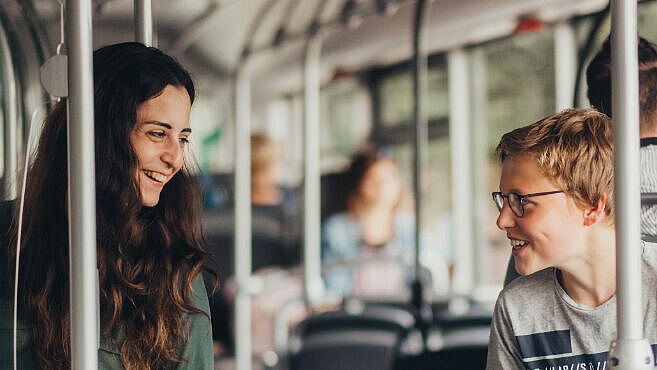 Jugendliche und Kind sitzen im Bus und lächeln sich über den Gang zu.