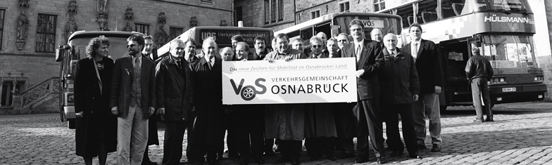 V O S Geschichte 1996. Schwarzweiß Fotografie zeigt eine Personengruppe mit einem Transparent für den Zusammenschluss zur V O S.