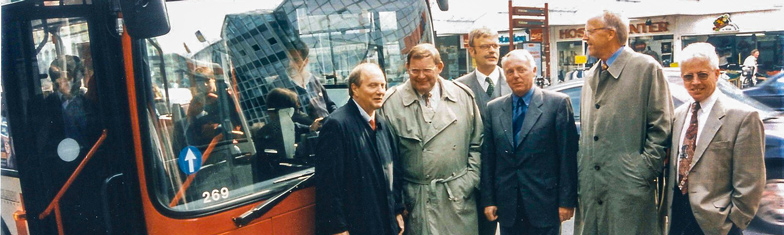 V O S Geschichte 1998. Fünf Herren in Mänteln und Anzügen stehen vor einem Linienbus.