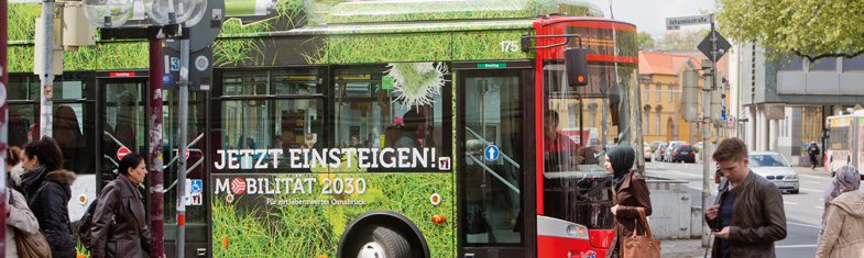 V O S Geschichte 2011. Linienbus im Stadtverkehr mit Werbung für grüne Mobilität und Fußgänger.